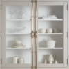 White kitchen cabinet with glass doors displaying stemware, bowls, and books on white shelves with brass handles left and right.