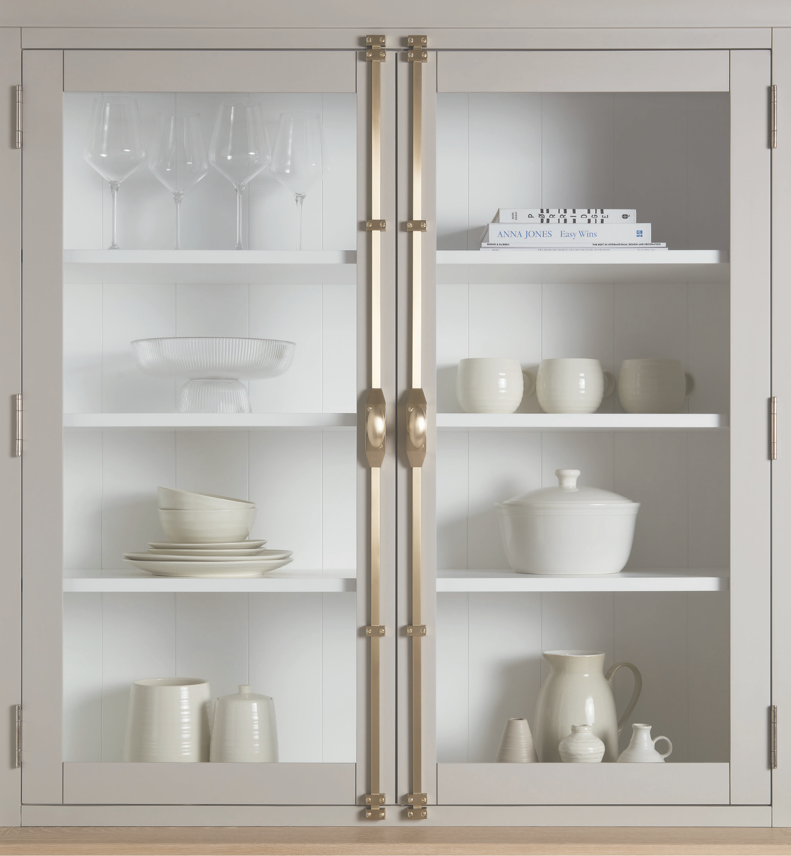 White kitchen cabinet with glass doors displaying stemware, bowls, and books on white shelves with brass handles left and right.